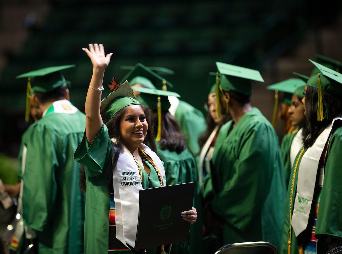 Student waving at UNT commencement wearing Hispanic Student Association stole
